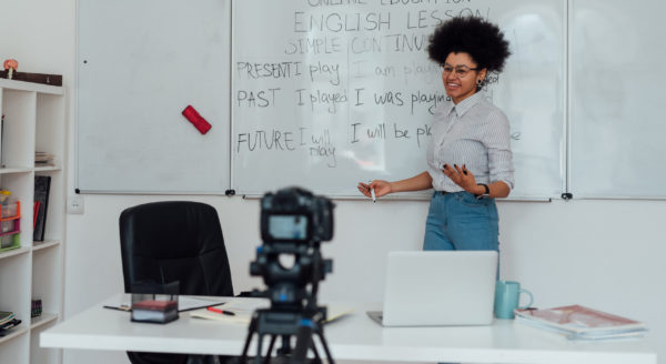 Corsi_online_Giving online class. Young afro american female English teacher standing near whiteboard and smiling, explaining rules of English grammar online. Main focus on woman. E-learning, distance education