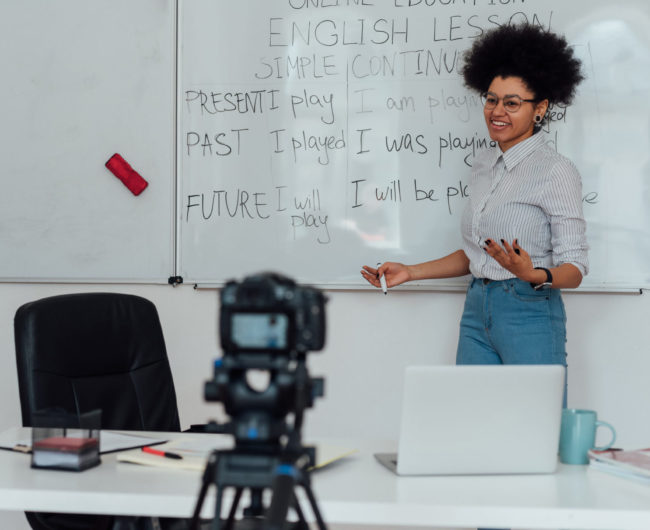 Corsi_online_Giving online class. Young afro american female English teacher standing near whiteboard and smiling, explaining rules of English grammar online. Main focus on woman. E-learning, distance education